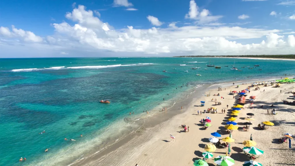 Praia de Maragogi com mar azul cristalino, faixa de areia clara, banhistas e barcos ao fundo.