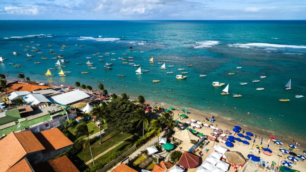 Vista aérea da praia de Porto de Galinhas com várias jangadas coloridas no mar e turistas na faixa de areia.