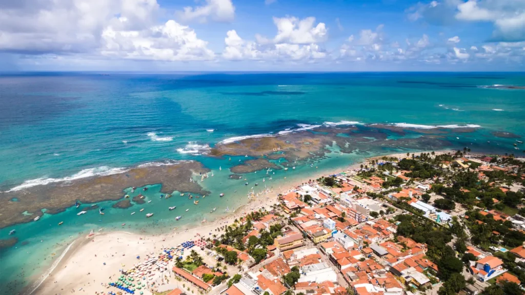 Vista aérea da praia de Porto de Galinhas maré com recifes expostos, mar azul e jangadas próximas às piscinas naturais.